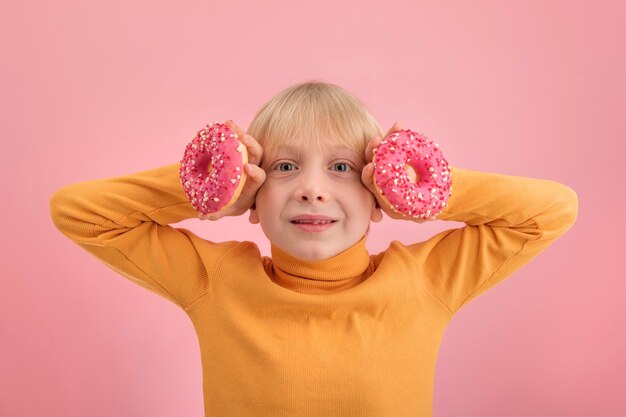 Portrait d'un garçon blond avec des beignets roses dans ses mains sur fond rose. Enfants et bonbons.
