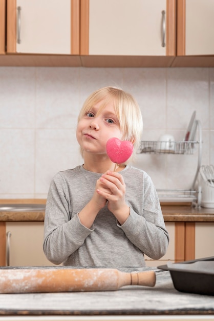 Portrait de garçon blond dans la cuisine avec une sucette en forme de coeur rose dans ses mains.