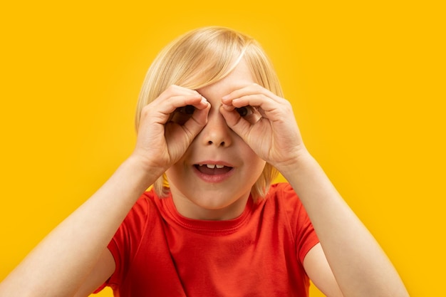 Portrait d'un garçon blond en T-shirt rouge gardant les mains comme des jumelles sur fond jaune