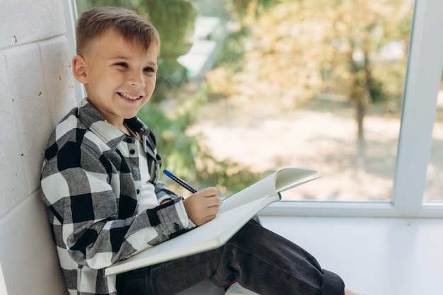 Portrait d'un garçon faisant ses devoirs Le garçon est assis sur le rebord de la fenêtre et écrit dans un cahier Un écolier fait ses devoirs près de la fenêtre Étude à domicile