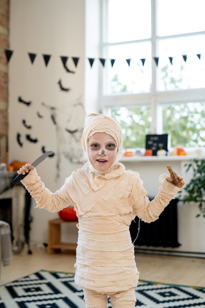 Portrait d'un garçon mignon en costume de momie debout avec un couteau et un biscuit comme symbole de la tradition des bonbons ou du sort pour Halloween