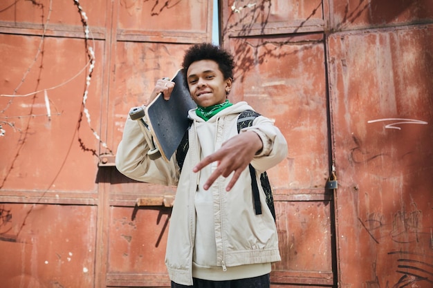 Portrait d'un garçon de patineur afro-américain tenant une planche à roulettes et faisant des gestes à la caméra en zone urbaine