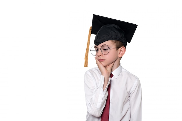 Portrait d'un garçon pensif dans une chemise blanche, des lunettes et un bonnet académique. Concept d'école. Isoler