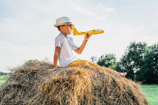 Portrait d'un garçon pieds nus au chapeau sur une botte de foin dans le champ. Jouer avec un avion jouet. Journée ensoleillée légère. Concept joyeux et musical. Activité de plein air. Vue de côté. Arbres en arrière-plan, Campagne