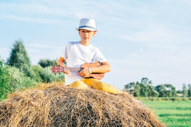 Portrait d'un garçon pieds nus au chapeau sur une botte de foin dans le champ. Jouer de la petite guitare, ukulélé. Journée ensoleillée légère. Concept joyeux et musical. Activité de plein air. Arbres en arrière-plan, Campagne