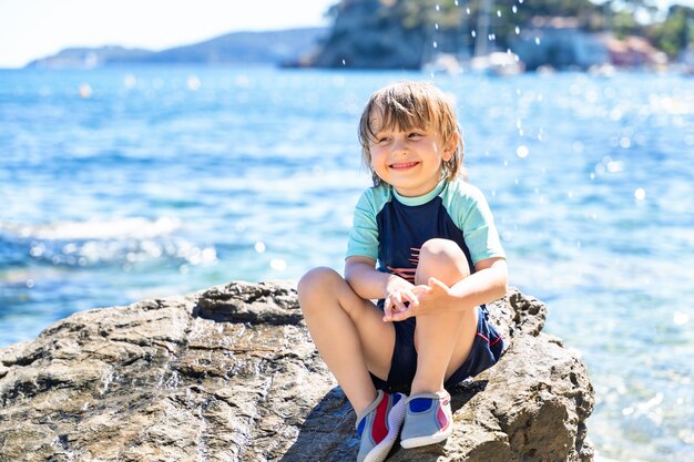 portrait d'un garçon souriant regardant ailleurs, assis sur un rocher au bord de la mer.