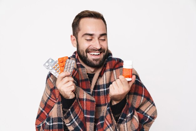 Portrait d'un gars caucasien heureux enveloppé dans une couverture tenant un tas de comprimés à cause de la grippe isolée sur un mur blanc