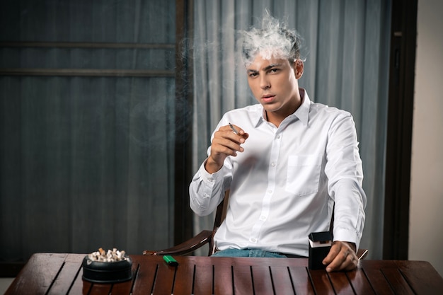 Portrait d'un gars posant assis à une table sur laquelle il y a un cendrier plein de cigarettes