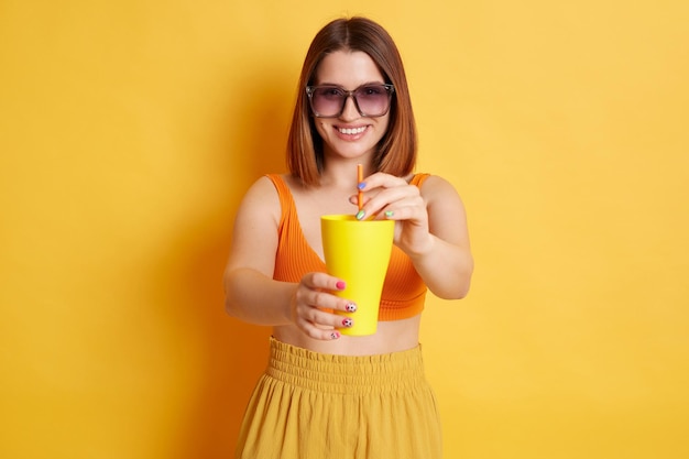 Portrait d'une gentille femme caucasienne souriante en lunettes de soleil portant des vêtements d'été posant isolé sur fond jaune vous offrant une boisson fraîche tenant une tasse avec une délicieuse boisson fraîche