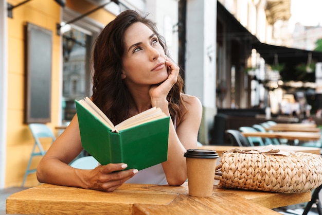 Portrait d'une gentille femme avec un sac de paille buvant du café dans une tasse en papier et lisant un livre assis dans un café confortable à l'extérieur