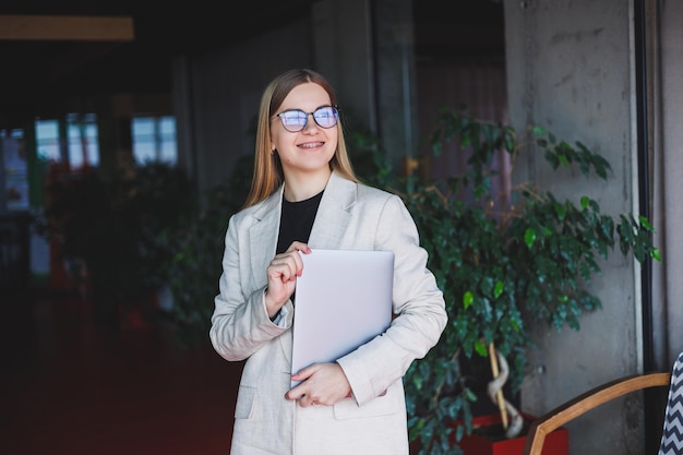 Portrait d'un gestionnaire prospère au bureau souriant pendant la journée de travail femme heureuse avec ordinateur portable profitant du temps dans un bureau moderne Femme aux cheveux blonds dans une veste beige et des lunettes