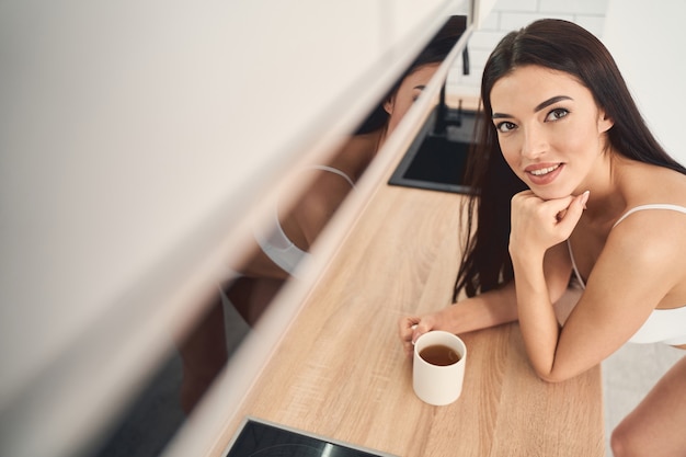 Portrait en grand angle d'une jeune femme souriante s'appuyant sur le plan de travail de la cuisine et levant avec un sourire sensuel