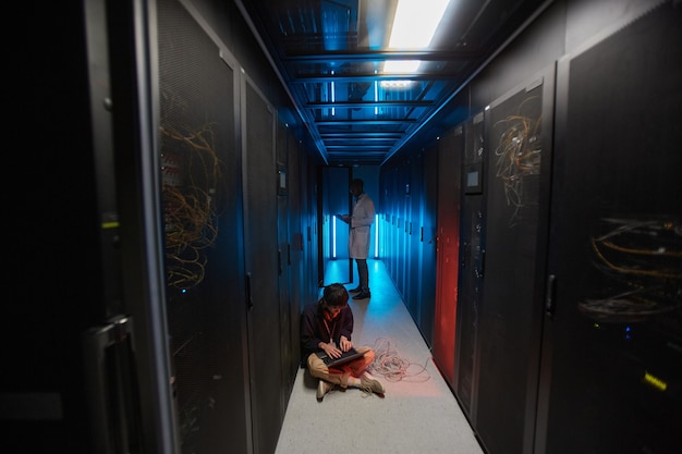 Portrait grand angle d'un jeune homme asiatique utilisant un ordinateur portable assis sur le sol dans la salle des serveurs éclairée par la lumière bleue et mettant en place un réseau de superordinateurs, espace de copie