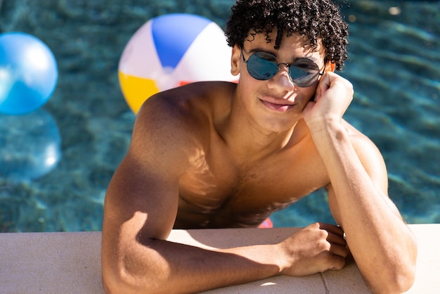 Portrait en grand angle d'un jeune homme torse nu biracial portant des lunettes de soleil penché au bord de la piscine. Inaltéré, beau, dreadlocks, style de vie, plaisir, concept d'activités d'été et de week-end.