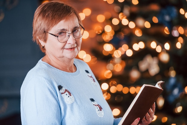 Portrait de grand-mère qui se tient dans la chambre décorée du nouvel an avec un livre.