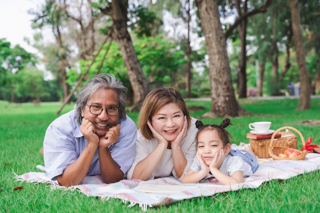 Portrait de grands-parents asiatiques et petite-fille portant sur le champ de verre vert en plein air, famille appréciant pique-nique ensemble dans le concept de la journée d&#39;été