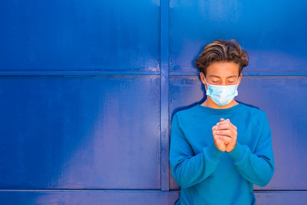 Portrait et gros plan d'un adolescent priant et portant un masque médical et chirurgical sur son visage - millénaire priant les yeux fermés portant un pull bleu et sur fond bleu