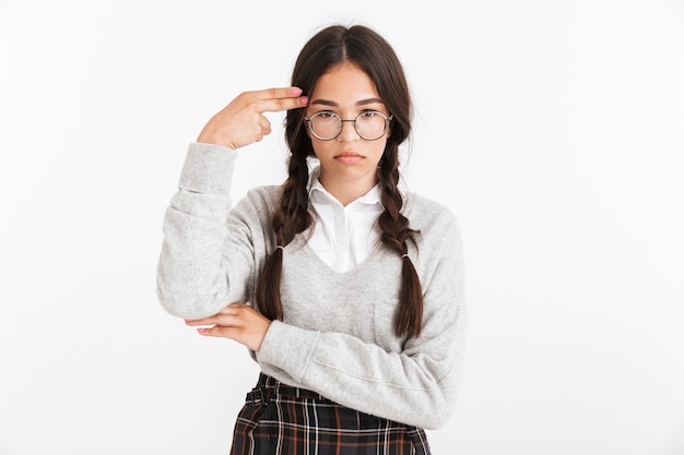 Portrait en gros plan d'une adolescente déprimée portant des lunettes et un uniforme scolaire montrant un geste de pistolet à doigt à son temple isolé sur un mur blanc