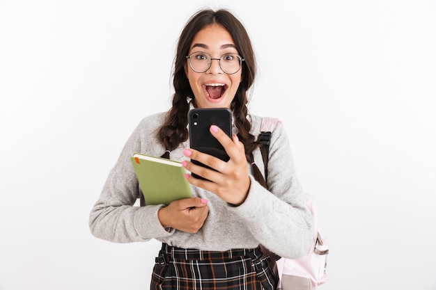 Portrait en gros plan d'une adolescente excitée portant des lunettes en riant tout en étudiant des livres et un smartphone isolé sur un mur blanc