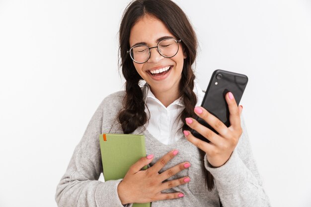 Portrait en gros plan d'une adolescente joyeuse portant des lunettes en riant tout en étudiant des livres et un smartphone isolé sur un mur blanc