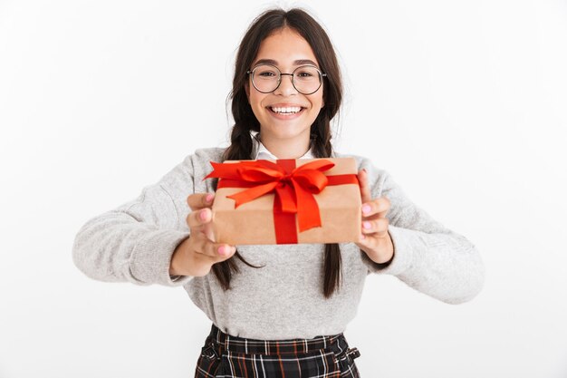 Portrait en gros plan d'une adolescente ravie portant des lunettes souriant tout en tenant une boîte présente avec un arc rouge isolé sur un mur blanc