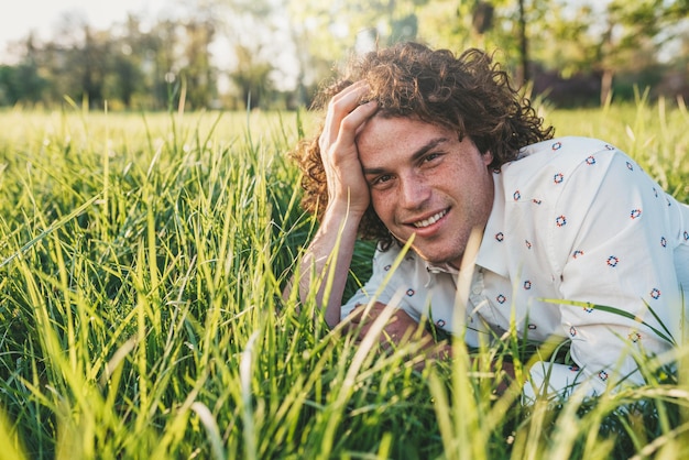 Portrait en gros plan d'un beau jeune homme aux cheveux bouclés souriant et relaxant sur l'herbe verte dans le parc en regardant la caméra Espace de copie pour la publicité Concept de personnes et de style de vie