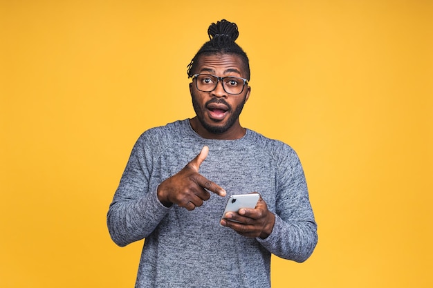 Portrait en gros plan d'un beau jeune homme noir afro-américain, choqué, surpris, bouche grande ouverte, fou de ce qu'il voit sur son téléphone portable, isolé sur fond jaune.