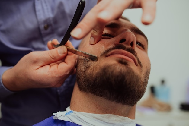 Portrait en gros plan d'un beau jeune homme se rasant la barbe avec un rasoir droit. Concentrez-vous sur la lame. Mise au point sélective. Photo de haute qualité