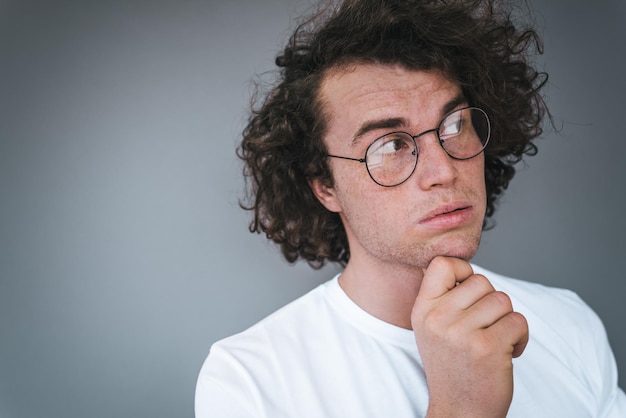 Portrait en gros plan d'un beau mâle pensif aux cheveux bouclés portant un t-shirt blanc et des lunettes rondes à la mode levant les yeux et pensant à des idées tenant son menton sur un mur gris Concept de personnes