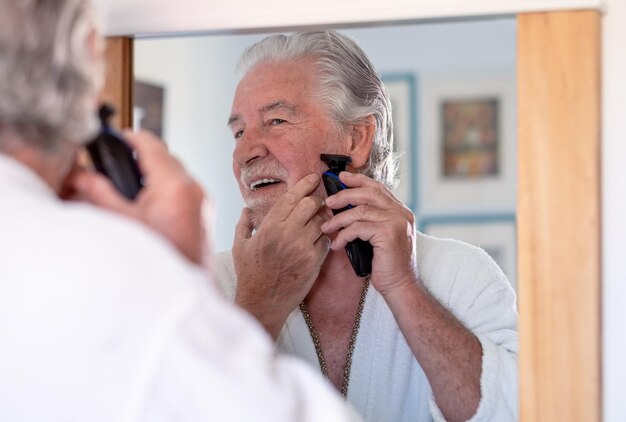 Portrait en gros plan d'un bel homme âgé en peignoir rasant la barbe avec un rasoir électrique