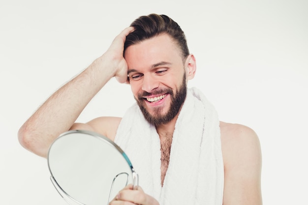 Portrait en gros plan d'un bel homme barbu avec une serviette regardant son reflet dans le miroir de la salle de bain isolé sur fond blanc