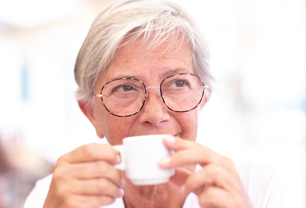 Portrait en gros plan d'une belle femme caucasienne âgée aux cheveux blancs et aux lunettes alors qu'elle regarde pensivement loin tout en buvant une tasse de café