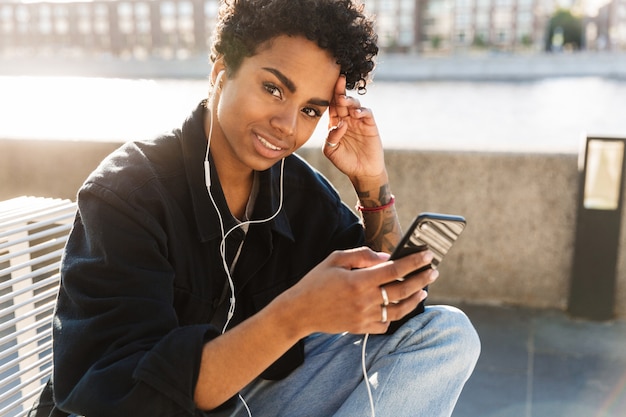 Photo portrait en gros plan d'une belle femme écoutant de la musique sur un téléphone portable avec des écouteurs assis sur un banc près de la rivière