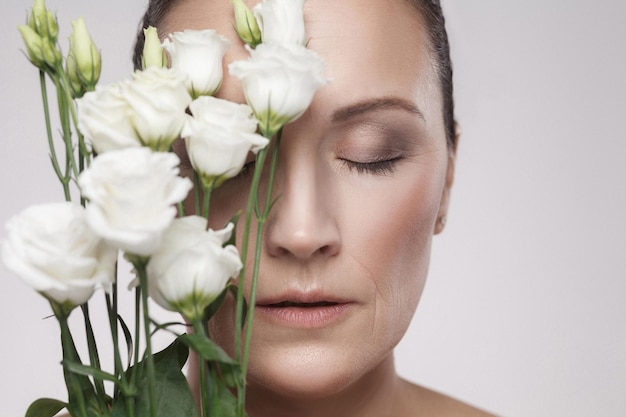 Portrait en gros plan d'une belle femme mature avec une peau parfaite et des rides âgées s'appuyant sur des fleurs. Concept de soins de la peau, cosmétiques vieillissants, isolés sur fond gris, espace pour copie, prise de vue en studio, intérieur