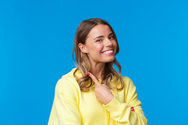 Portrait de gros plan de la belle fille blonde aux cheveux courts pointant sur son piercing à l'oreille, souriant fier et satisfait, change son look, debout sur un mur bleu en sweat à capuche jaune