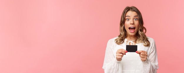 Portrait en gros plan d'une belle fille blonde émotive, excitée et ravie, montrant une carte de crédit et se sentant très heureuse de parler de remises offres spéciales service bancaire fond rose