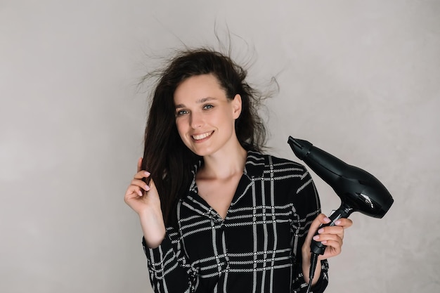 Portrait en gros plan d'une belle fille brune en pyjama noir séchant la coiffure et souriant