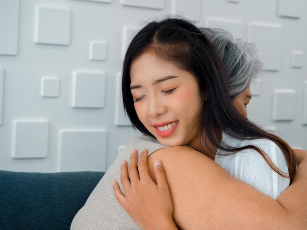 Portrait en gros plan d'une belle fille ou d'un petit-enfant asiatique heureux embrassant sa mère aînée ou ses grands-parents aux cheveux blancs avec soin et confort tout en étant assis sur un canapé gris dans le salon à la maison