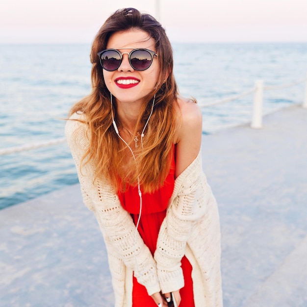 Portrait en gros plan d'une belle fille en robe rouge et veste blanche sur une jetée, souriant et écoutant de la musique sur des écouteurs sur un smartphone. Elle porte des lunettes de soleil foncées.