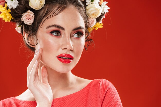 Portrait en gros plan d'une belle jeune femme brune avec une coiffure florale et un maquillage lumineux posant isolé sur un mur rouge, en détournant les yeux