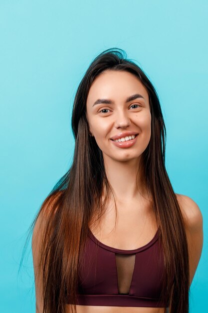 Portrait en gros plan d'une belle jeune femme brune en tenue de sport souriante sur fond bleu.