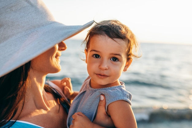 Portrait en gros plan d'une belle mère heureuse étreint sa jolie fille portant un chapeau blanc et une robe rayée posant à la mer Maman avec une petite fille marchant à l'océan Femme avec bébé posant à l'océan