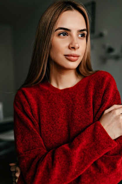 Portrait en gros plan d'une charmante dame avec un maquillage nude et de longs cheveux clairs portant un pull rouge et regardant de côté avec un sourire heureux