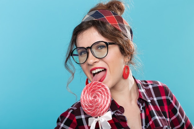 Portrait en gros plan d'une charmante jeune femme pin-up dans une chemise à carreaux et des lunettes lèche une grosse sucette contre une surface bleue