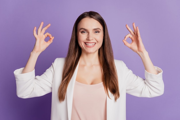 Portrait en gros plan d'une dame montrant deux signes okey portant un costume formel posant sur un mur violet