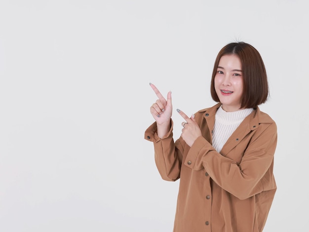 Portrait en gros plan de la découpe en studio d'un jeune mannequin asiatique aux cheveux courts et aux cheveux courts en veste de manteau marron debout souriant regardant la caméra pointant le doigt vers le haut sur l'espace de copie vierge sur fond blanc.