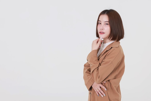 Portrait en gros plan de la découpe en studio d'un jeune mannequin asiatique aux cheveux courts et aux cheveux courts en veste de manteau marron debout souriant regardant la caméra pointant le doigt vers le haut sur l'espace de copie vierge sur fond blanc.