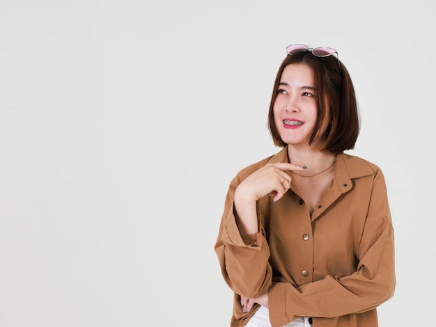 Portrait en gros plan de la découpe en studio d'un jeune mannequin asiatique aux cheveux courts et aux cheveux courts en veste de manteau marron debout souriant regardant la caméra pointant le doigt vers le haut sur l'espace de copie vierge sur fond blanc.