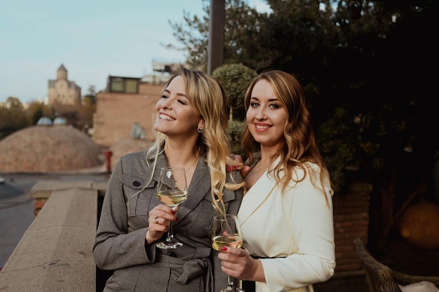 Portrait en gros plan de deux amies en costumes stricts riant en buvant du vin sur la terrasse à l'extérieur