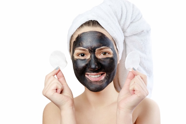 Portrait en gros plan drôle belle femme avec masque facial noir sur fond blanc, fille avec une serviette blanche sur la tête, sourire satisfait et heureux, tient dans les mains les cotons pour se démaquiller